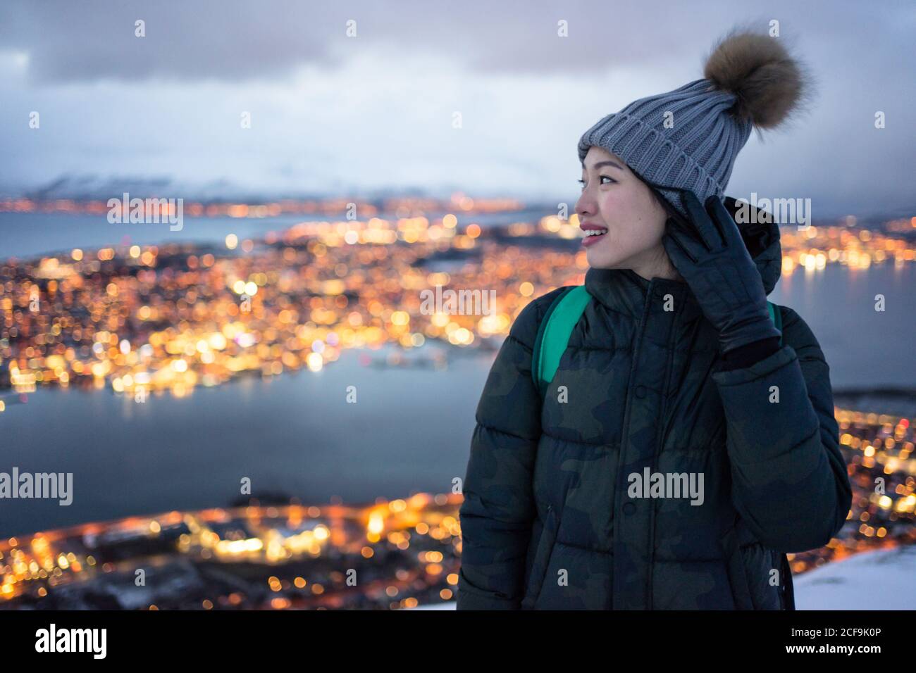 Jeune femme excitée en veste en duvet kaki et gris chaud chapeau regardant loin et contemplant la vue d'hiver incroyable de la ville situé sur la côte en soirée Banque D'Images