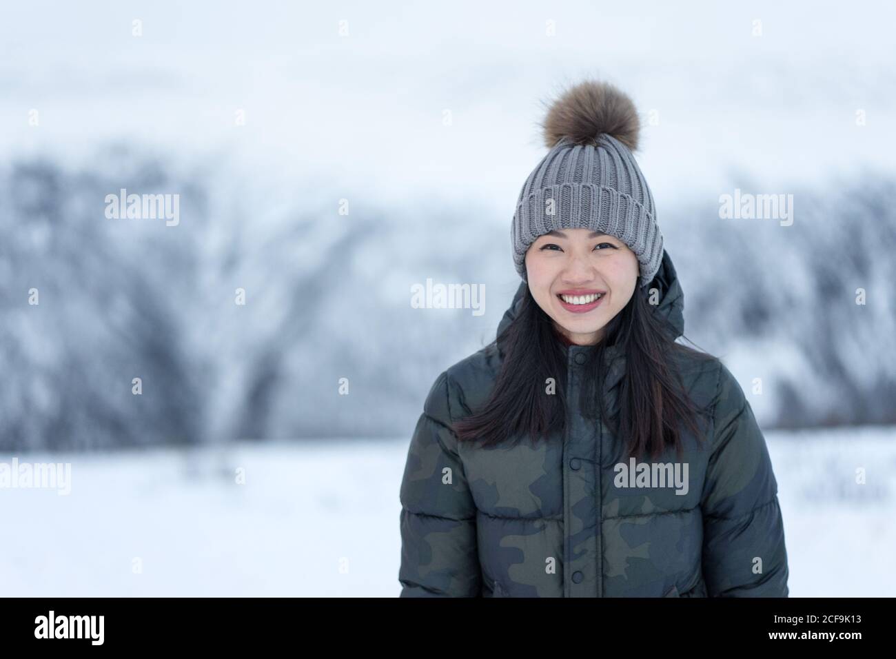 Magnifique femme heureuse avec des yeux étonnants dans le noir repéré vers le bas veste et chapeau chaud gris regardant l'appareil photo et souriant contre les paysages flous du parc enneigé en journée Banque D'Images