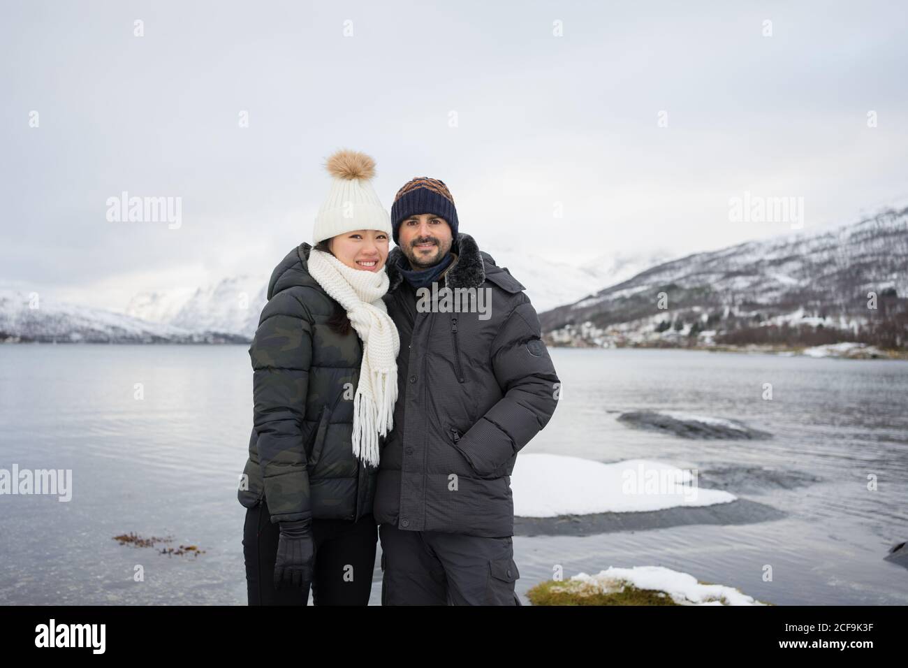 Joyeux couple dans la veste de duvet chaude et les chapeaux embrassant et sourire à l'appareil photo tout en se tenant sur la plage contre blanc et Collines enneigées brunes et eaux calmes des cols du lac Kaldfjorden En Norvège Banque D'Images