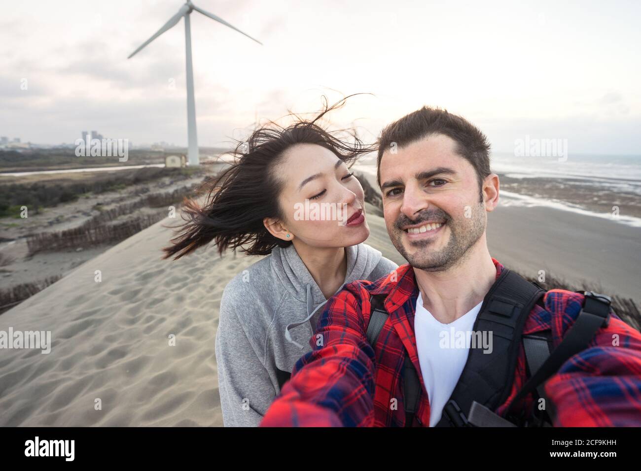 Joyeux couple multiracial dans une tenue décontractée cuddling tout en prenant le selfie et se tenir sur le sable près du bord de mer pendant les déplacements dans un ciel nuageux ciel et regarder la caméra Banque D'Images