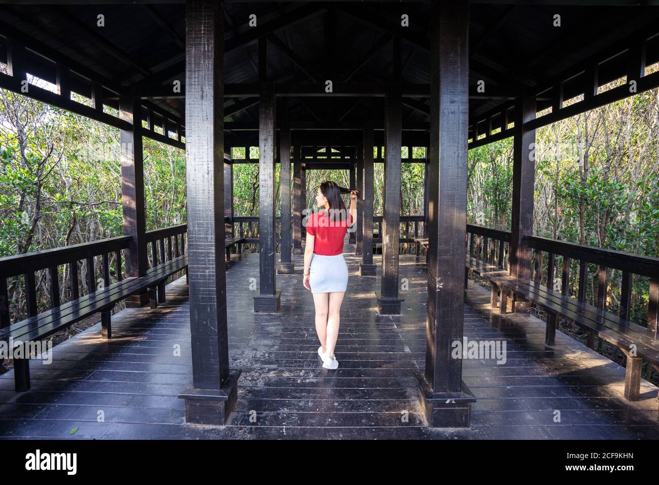 Vue arrière de la femme asiatique mince en jupe courte et t-shirt rouge marchant sur une ancienne terrasse en bois avec colonnes et des bancs entourés d'une forêt avec des arbres verts Banque D'Images