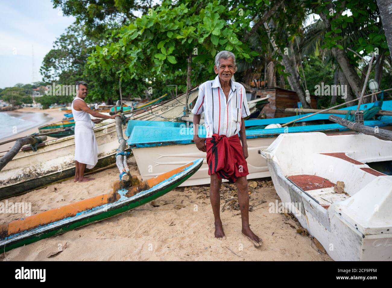 Tangalle, Sri Lanka - 28 juillet 2019 : pêcheur ethnique âgé à cheveux gris en vêtements légers décontractés debout sur un rivage sablonneux près des bateaux et regardant la caméra Banque D'Images