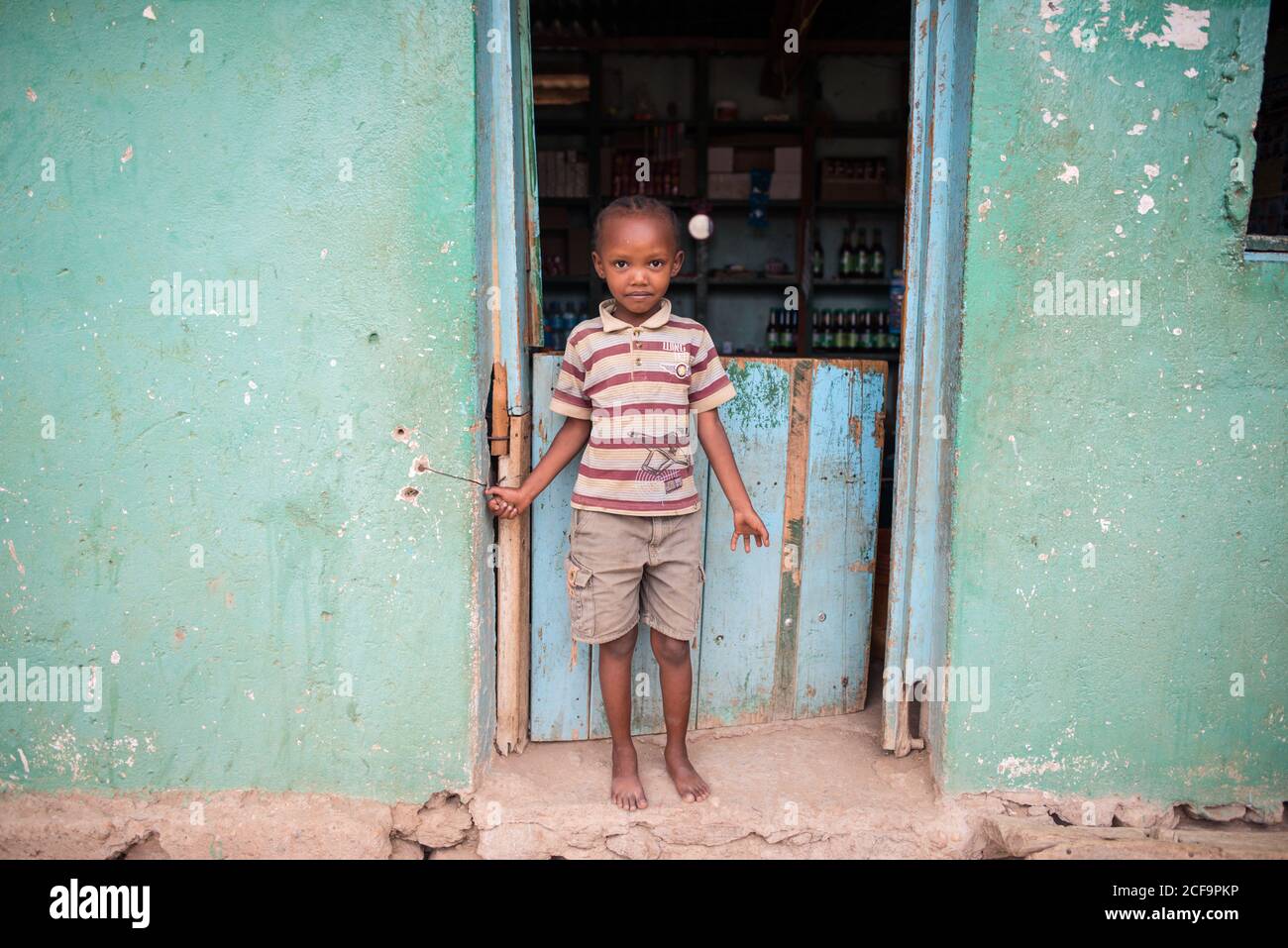 Tanzanie, Afrique de l'est - novembre, 2016: Petit garçon africain dans des vêtements décontractés debout et regardant la caméra près de la petite maison ancienne à la rue de la ville de Tanzanie Banque D'Images