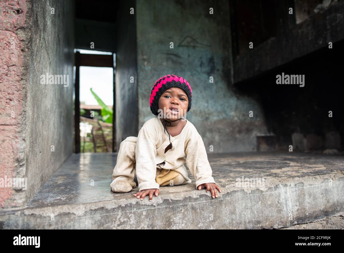 Ouganda - novembre, 26 2016: Joli bébé africain dans un pyjama sale et un  chapeau tricoté regardant la caméra tout en étant assis à l'intérieur d'un  bâtiment en béton non achevé Photo