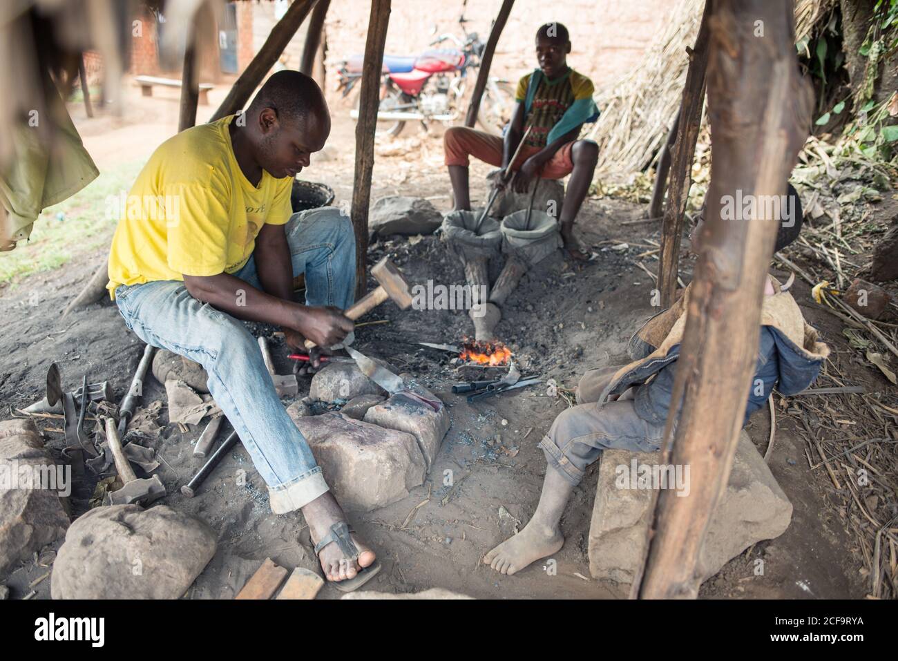 Ouganda - 26 novembre 2016 : forgeron africain adulte attentif dans des vêtements vifs et décontractés se concentrant et travaillant avec le métal en utilisant un gros marteau tout en étant assis à côté d'un jeune collègue avec des soufflets et des montres feu sous le toit de paille dans la forge rustique locale Banque D'Images