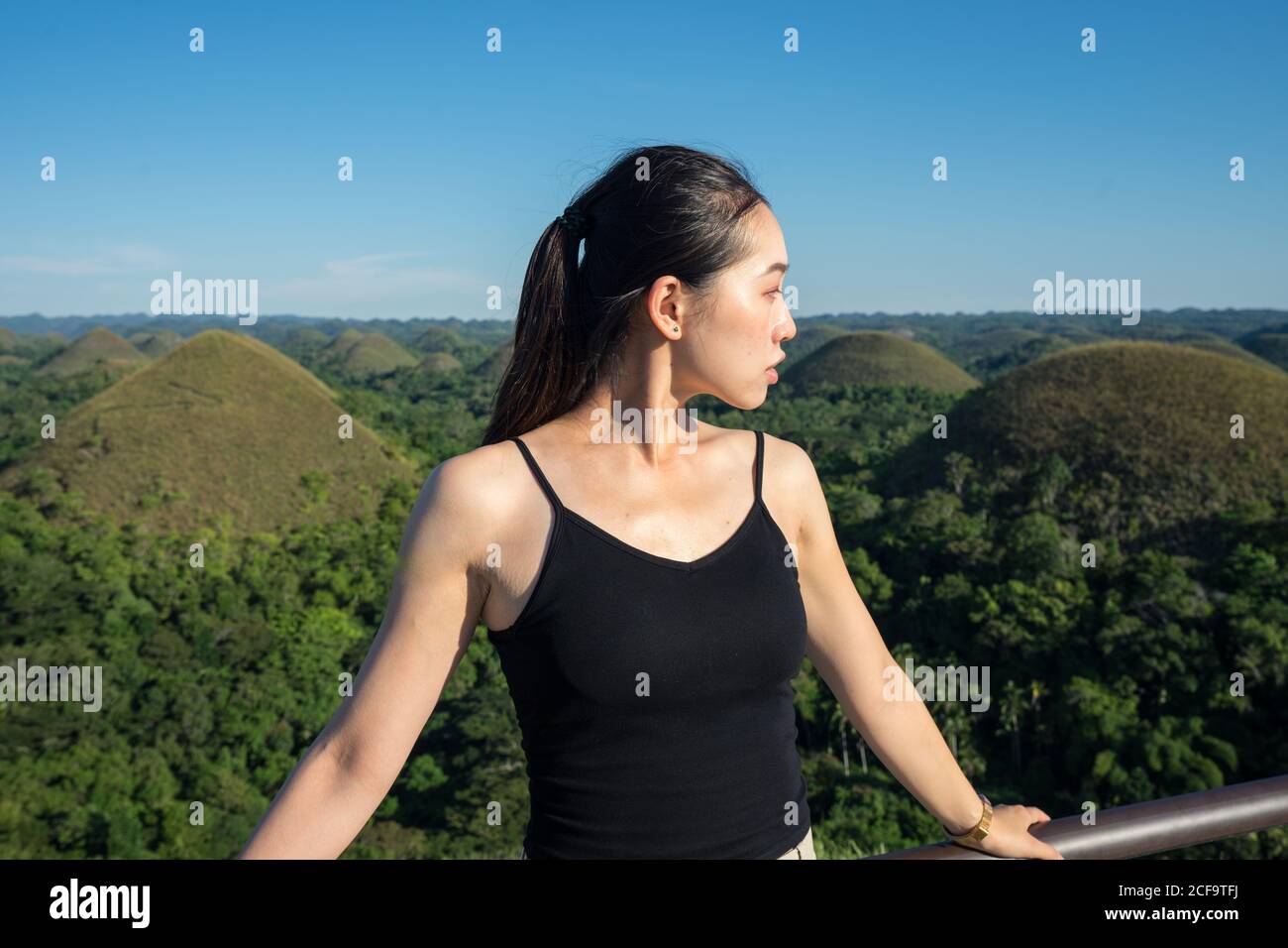 Jeune asiatique touriste en haut noir et pantalon gris vue sur l'extérieur en se tenant près de la clôture sur la terrasse d'observation avec des collines vertes et un ciel bleu en arrière-plan Banque D'Images