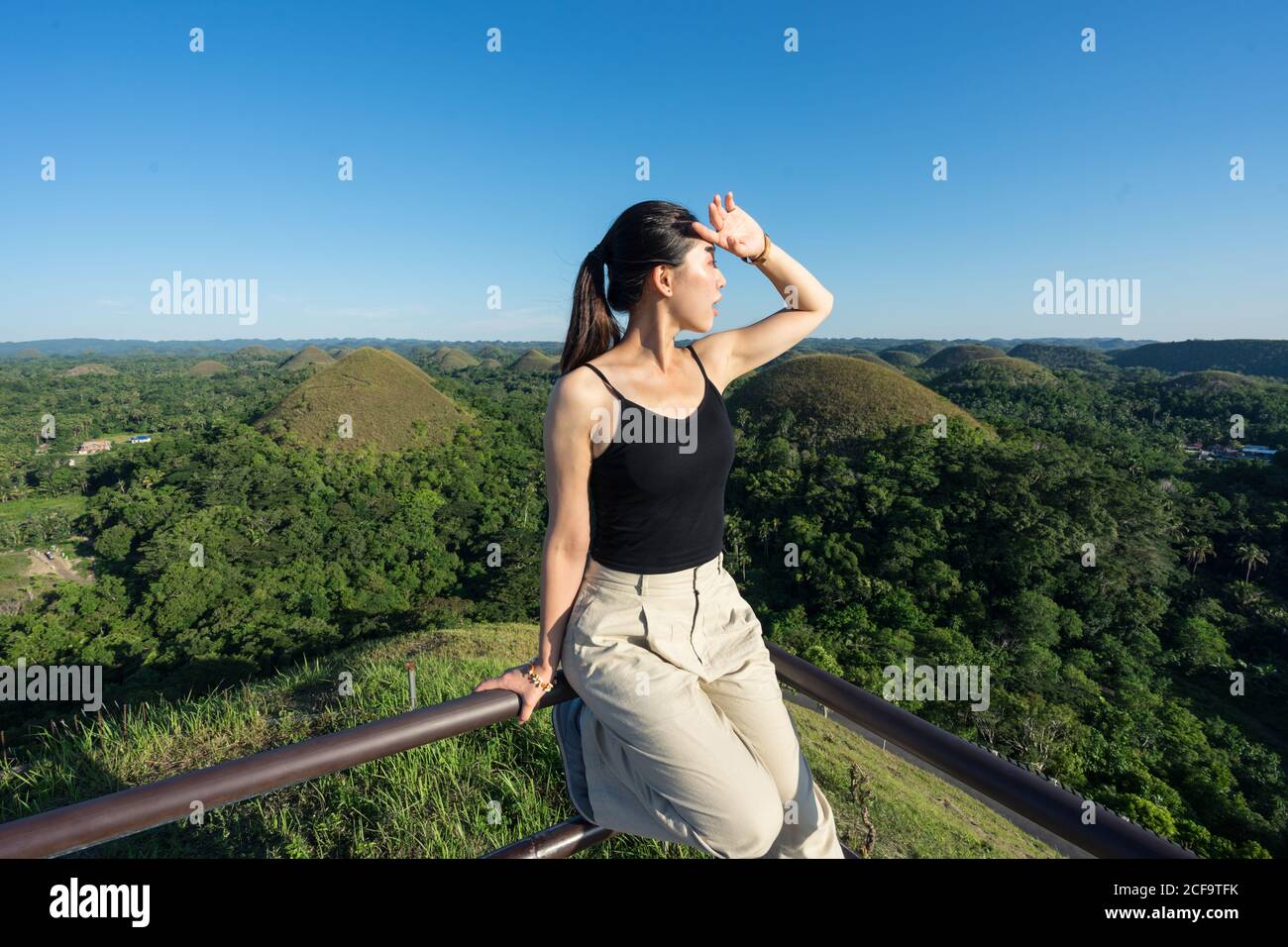 Joyeuse jeune asiatique touriste en noir et gris pantalon regardant loin et utilisant la main pour se cacher pour couvrir faites face au soleil tout en vous tenant à côté de la clôture en observation terrasse avec collines vertes et ciel bleu en arrière-plan Banque D'Images