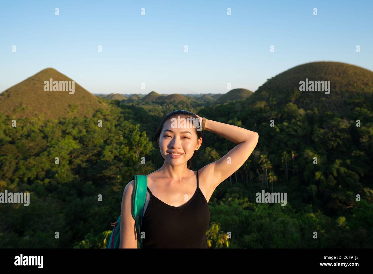 Joyeuse jeune asiatique touriste en noir et gris pantalon regardant la caméra à côté de la clôture en observation terrasse avec collines vertes et ciel bleu en arrière-plan Banque D'Images