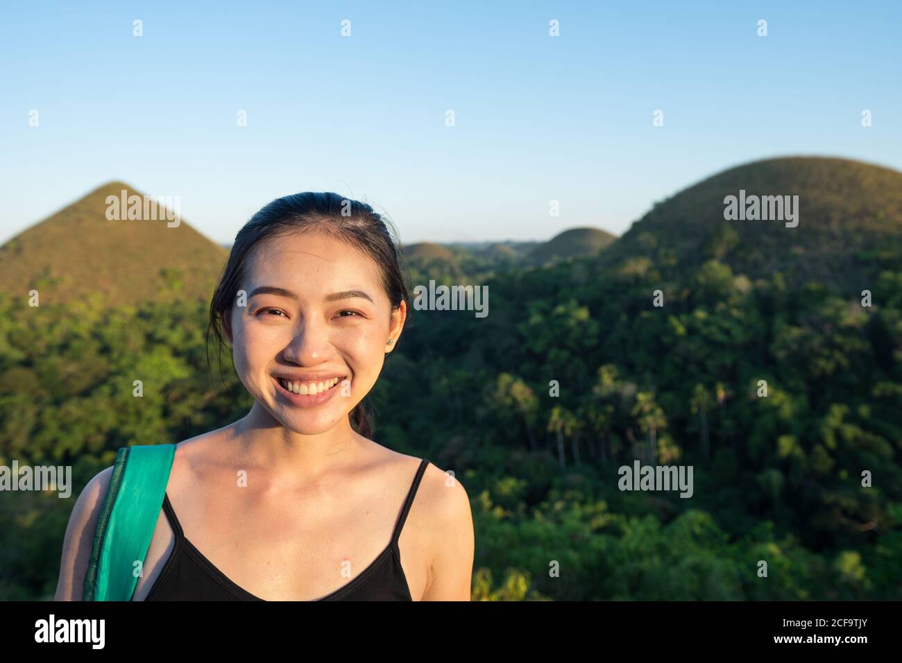 Joyeuse jeune asiatique touriste en noir et gris pantalon regardant la caméra à côté de la clôture en observation terrasse avec collines vertes et ciel bleu en arrière-plan Banque D'Images