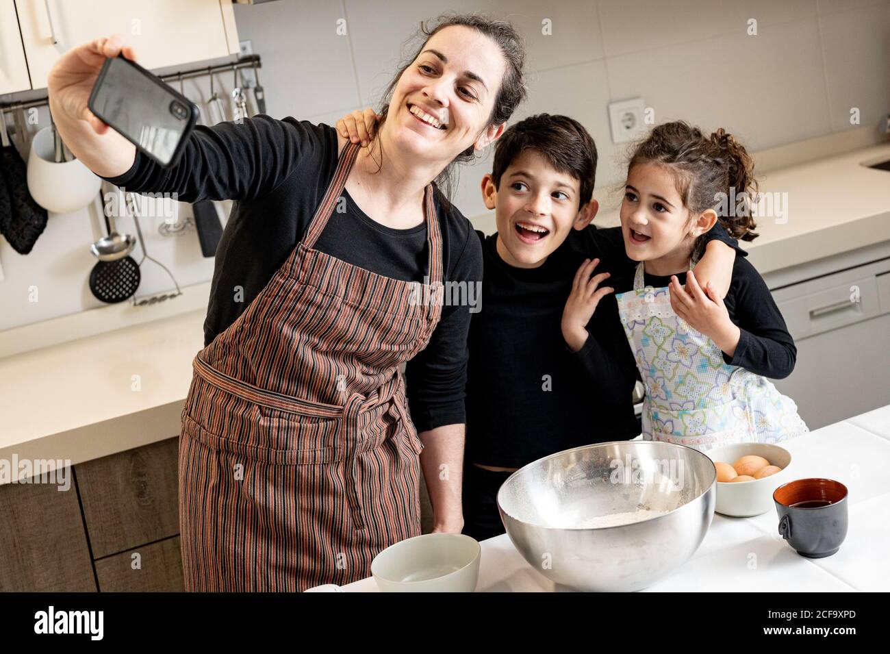 Une femme heureuse en tablier souriant et prenant le selfie avec un téléphone portable avec des enfants heureux tout en cuisinant des pâtisseries ensemble dans la cuisine confortable à la maison Banque D'Images