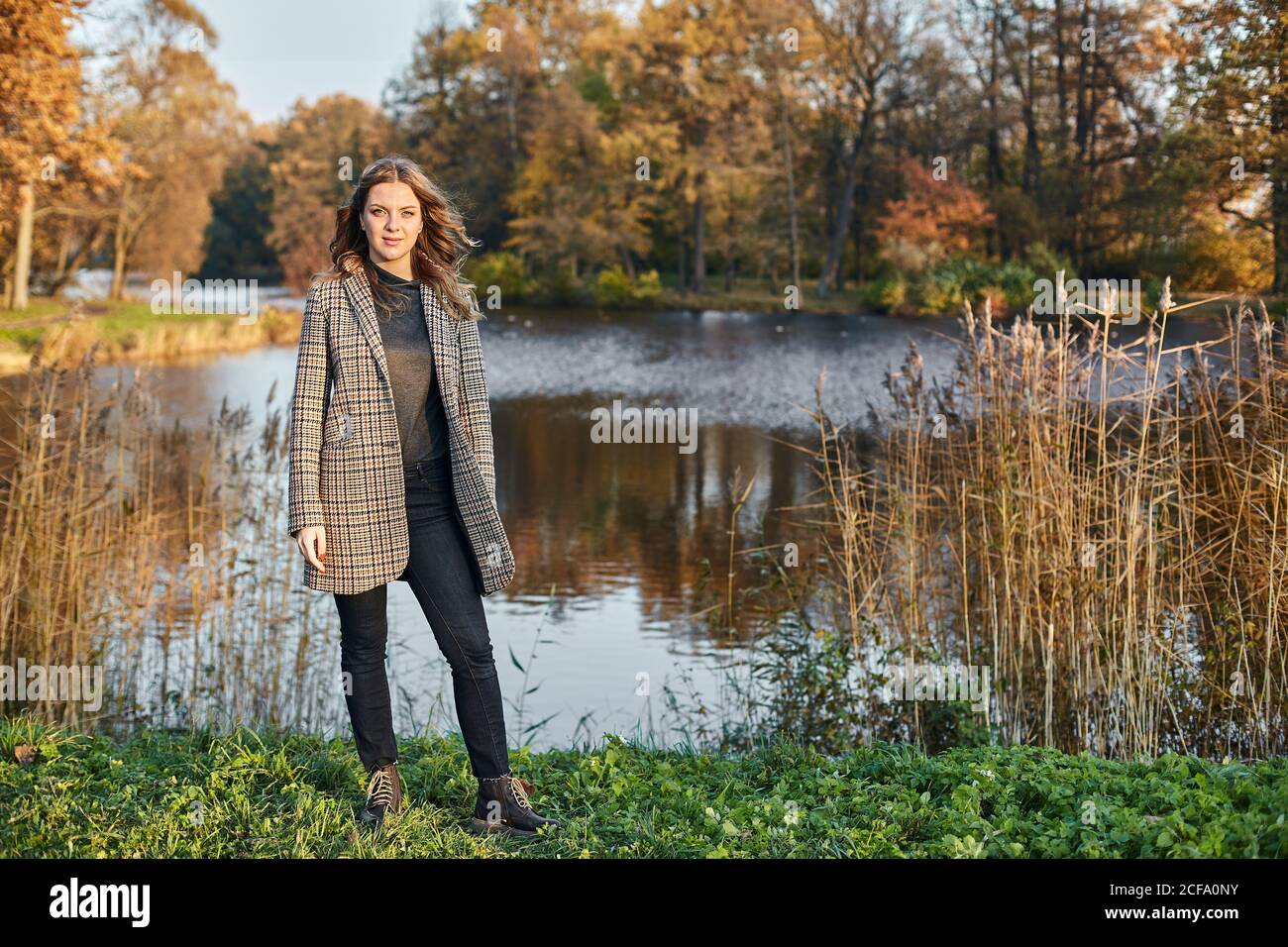 Femme souriante aux cheveux longs portant un manteau brun et debout à l'automne parc à un lac Banque D'Images