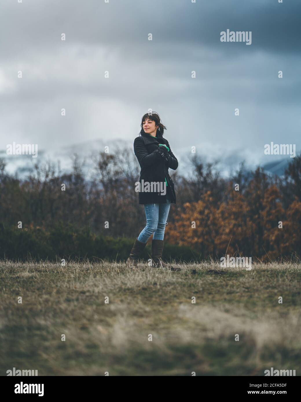 De dessous de jolie femme décontractée en manteau noir et jeans aux bras croisés et sensation de froid à l'automne champ avec collines couvertes de neige et ciel gris avec nuages sombres sur l'arrière-plan Banque D'Images