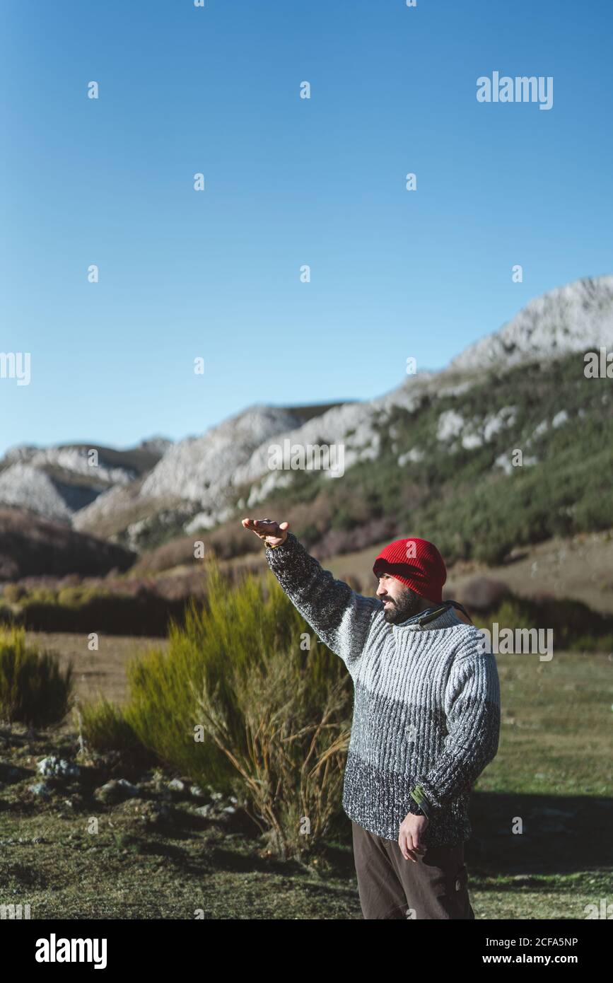 Vue latérale d'un adulte mâle barbu en chandail gris et casquette tricotée rouge debout avec main levée sur la montagne vallée et vue sur la distance le jour ensoleillé avec bleu ciel Banque D'Images