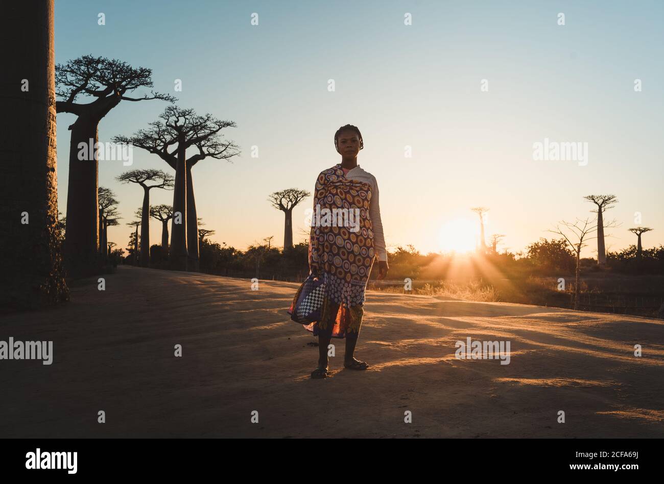 Madagascar - 6 JUILLET 2019: Femme ethnique confiante en tenue exotique multicolore debout en pente par de grands baobabs en rayons du soleil au crépuscule Banque D'Images