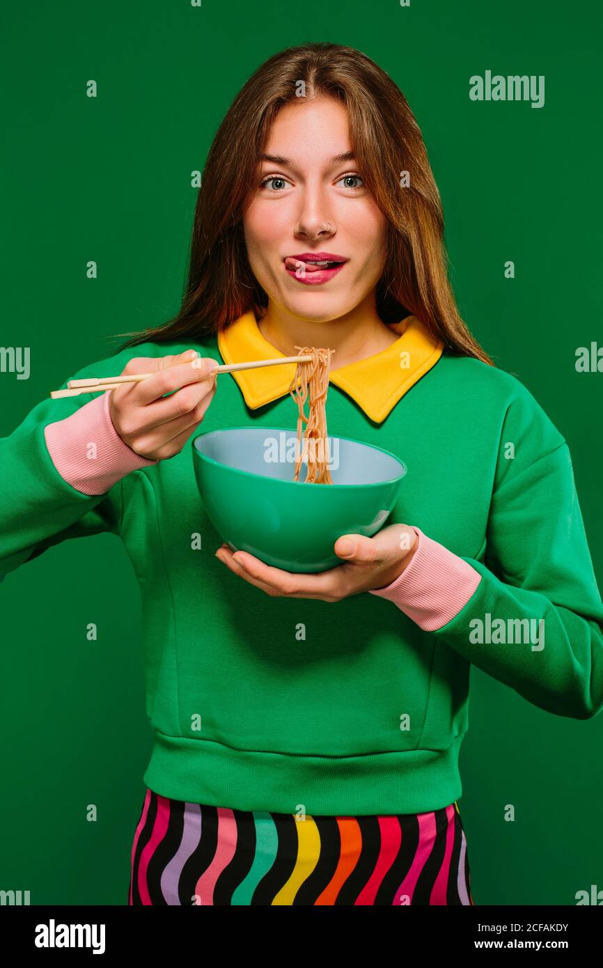 Jeune femme positive en chemise verte regardant l'appareil photo grimacing en collant la langue tout en mangeant de délicieuses nouilles instantanées avec des baguettes sur fond vert Banque D'Images