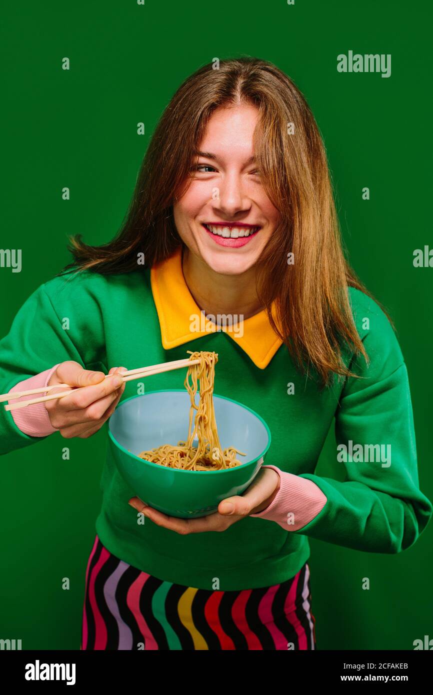Positive gai jeune femme en vert chemise regardant l'appareil photo tout en mangeant de délicieuses nouilles instantanées avec des baguettes sur fond vert Banque D'Images