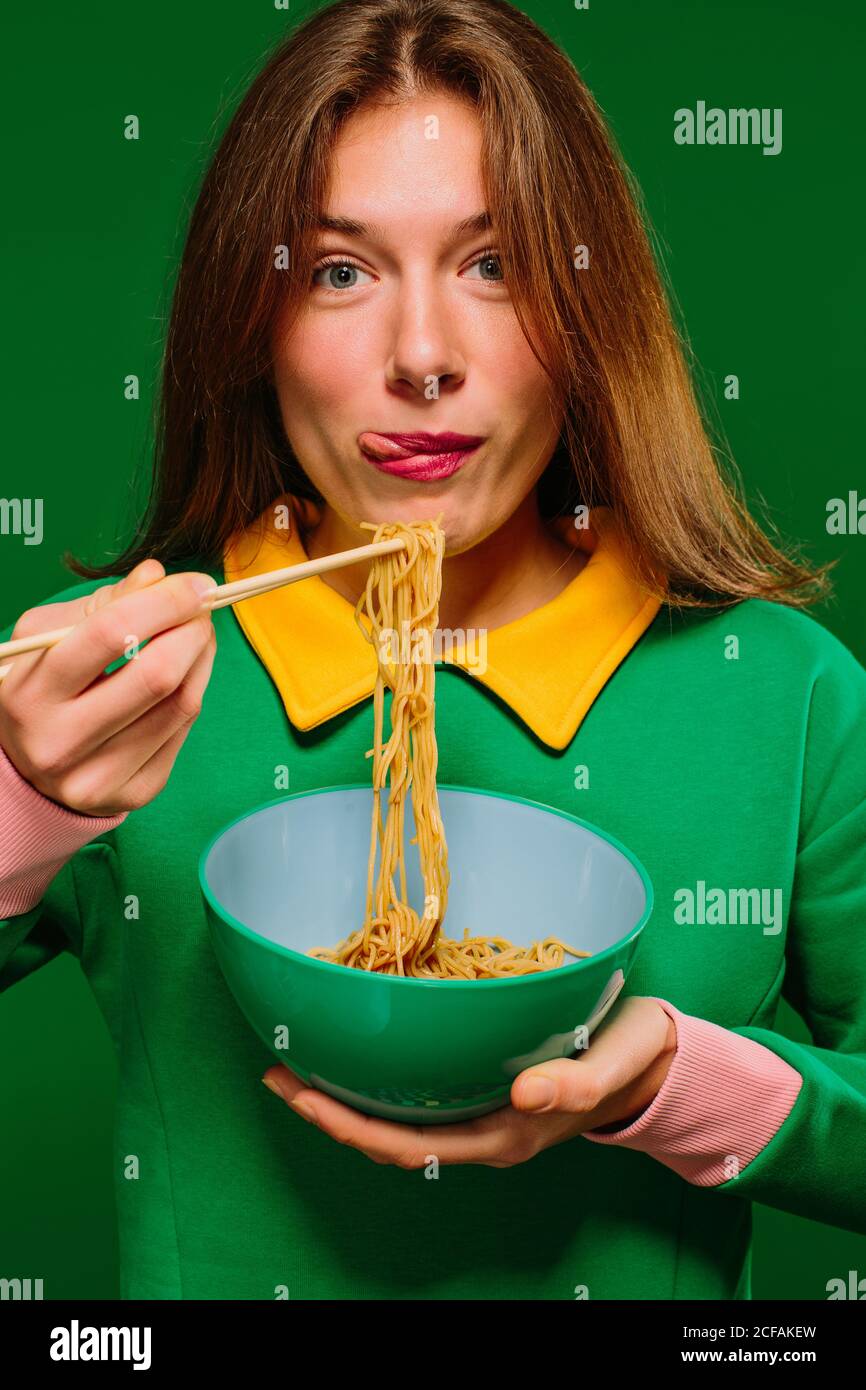 Jeune femme positive en chemise verte regardant l'appareil photo grimacing en collant la langue tout en mangeant de délicieuses nouilles instantanées avec des baguettes sur fond vert Banque D'Images