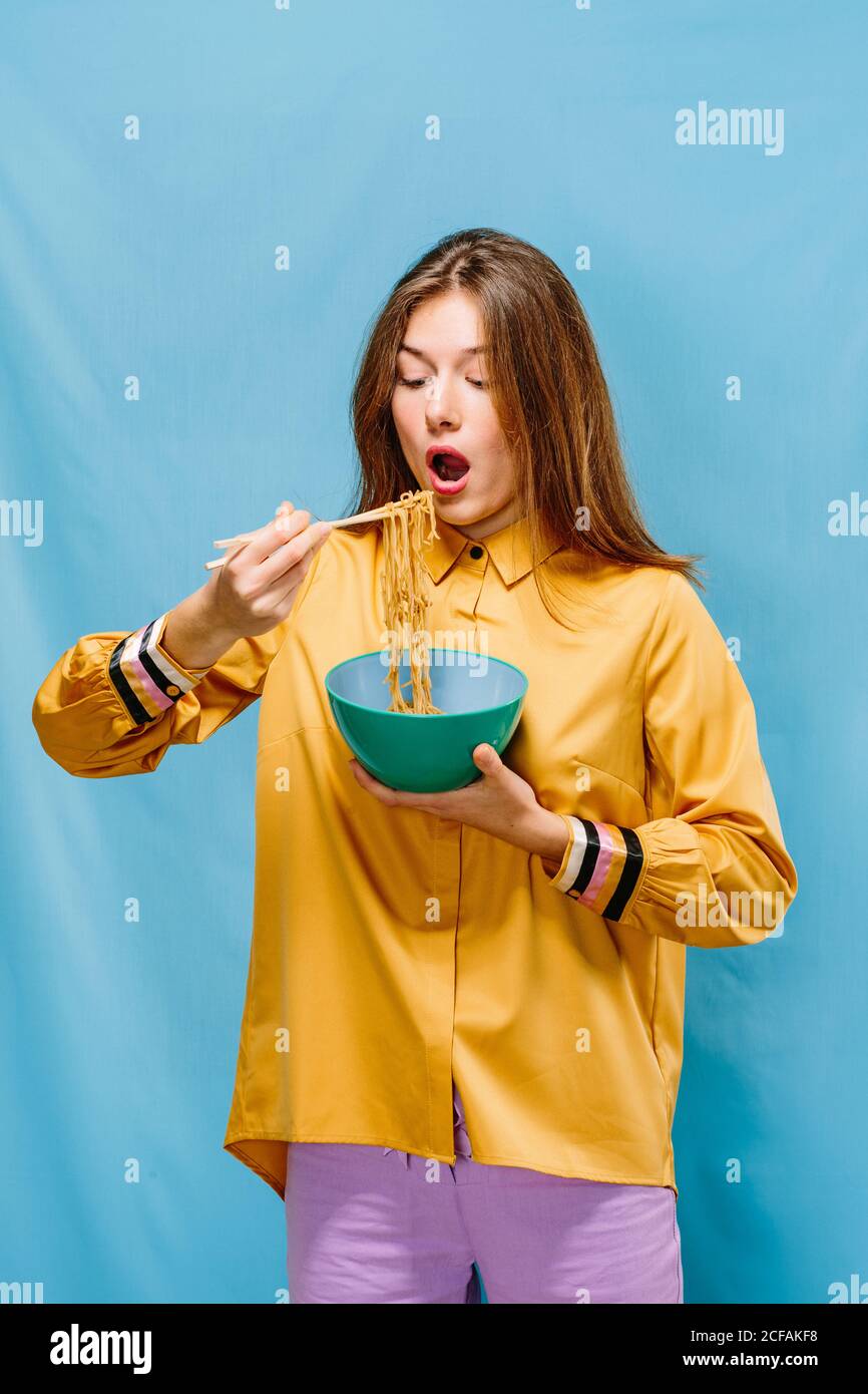 Positive jeune femme en chemise jaune ouvrant la bouche à manger nouilles instantanées délicieuses avec baguettes sur fond bleu Banque D'Images
