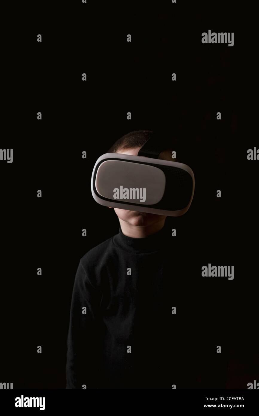 Enfant portant un chandail noir et faisant l'expérience d'un casque VR debout contre le mur noir Banque D'Images