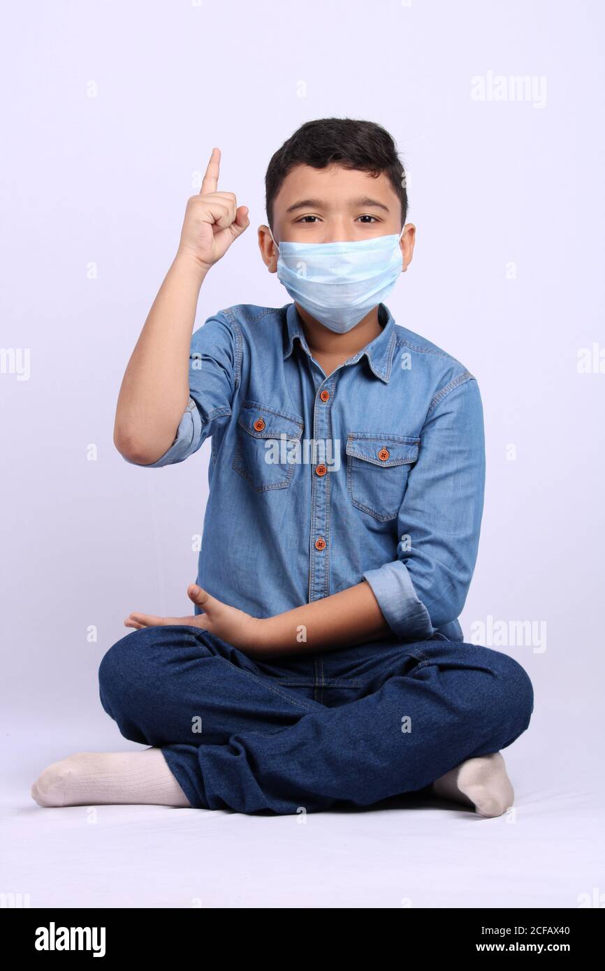 jeune garçon indien mignon portant un masque chirurgical pointant le doigt vers le haut tout en étant assis sur le sol. Banque D'Images
