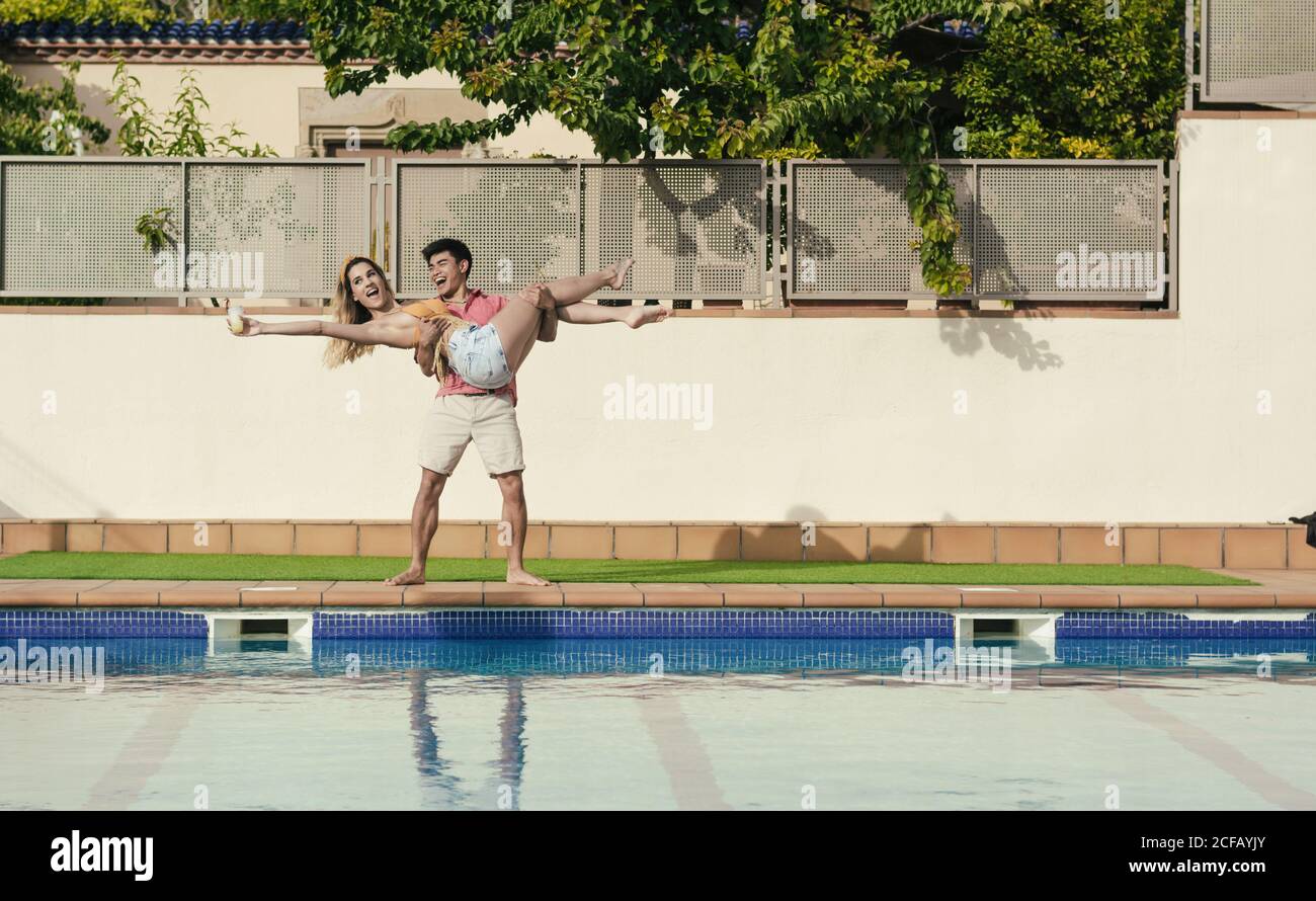 Groupe d'amis qui ont une fête au bord de la piscine pendant qu'ils dansent, rient et boivent des cocktails Banque D'Images