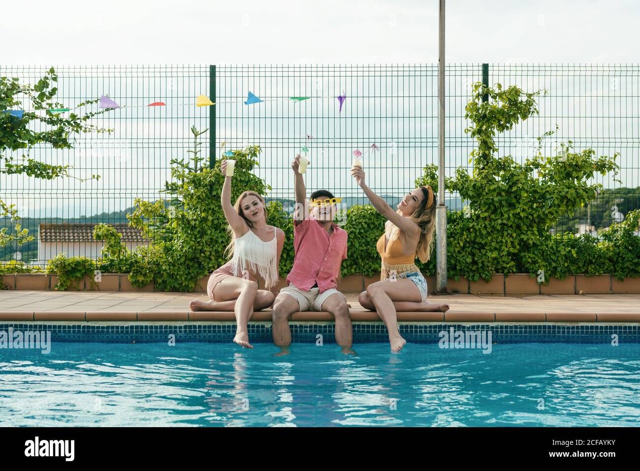 Groupe d'amis qui ont une fête au bord de la piscine pendant qu'ils dansent, rient et boivent des cocktails Banque D'Images
