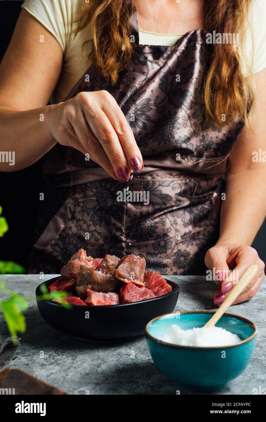 Femme sans visage portant un tablier debout à la table et ajoutant du sel dans un bol avec des tranches de bœuf cru tout en préparant la viande pour la cuisson Banque D'Images