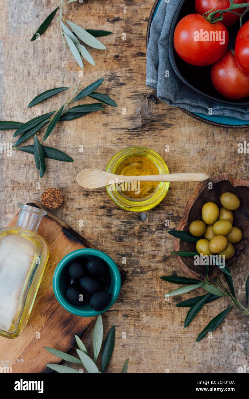 Du dessus du pot en verre avec de l'huile d'olive biologique avec cuillère en bois placée sur une table grise avec des tomates fraîches olives vertes et noires près des branches d'arbres Banque D'Images