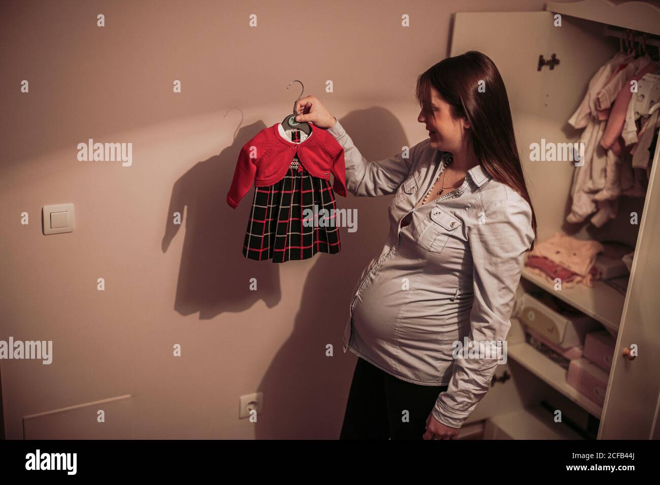 D En Haut Vue Laterale De La Jeune Femme Heureuse S Attendant A Bebe Debout Dans La Chambre De Bebe Rose A Cote De L Armoire Ouverte Et Tenant Dans Un Cintre A Main Avec