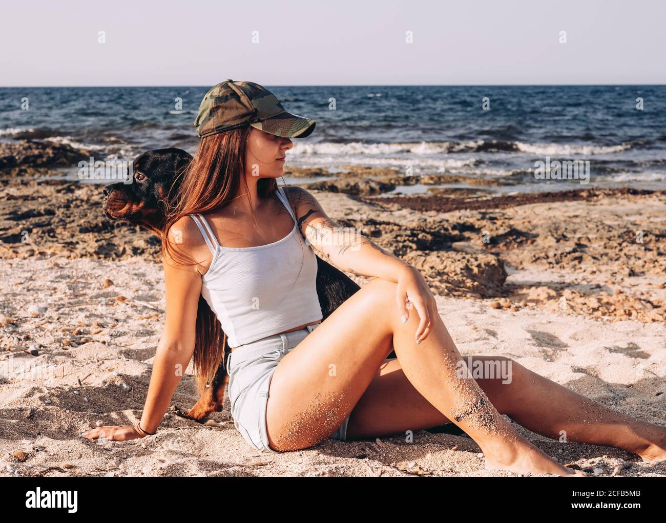 Une jeune femme positive dans des vêtements décontractés et une casquette  Plage de sable avec le chien de Rottweiler tout en passant du temps au  soleil jour en été Photo Stock -