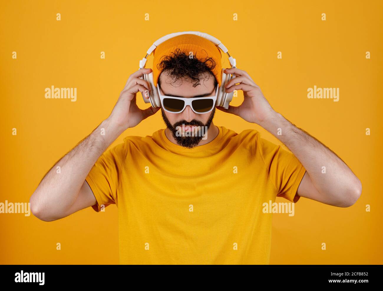 Un homme barbu dans une tenue élégante écoutant de la musique sur un casque blanc contemporain sur fond jaune Banque D'Images