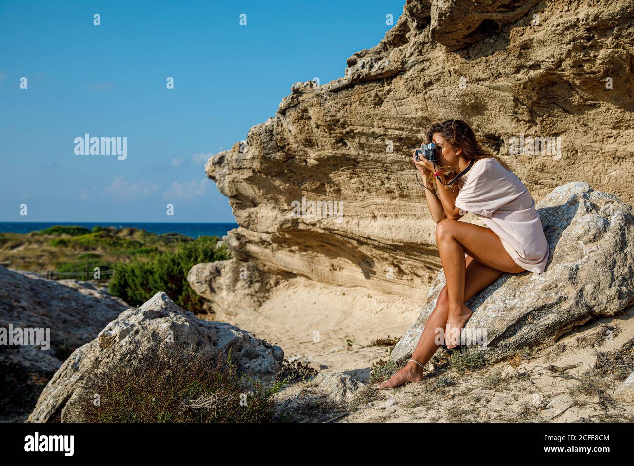 Vue latérale d'une femme cintrée bronzée en chemise blanche prenant des photos confortablement placée sur un rocher pierreux par temps lumineux Banque D'Images