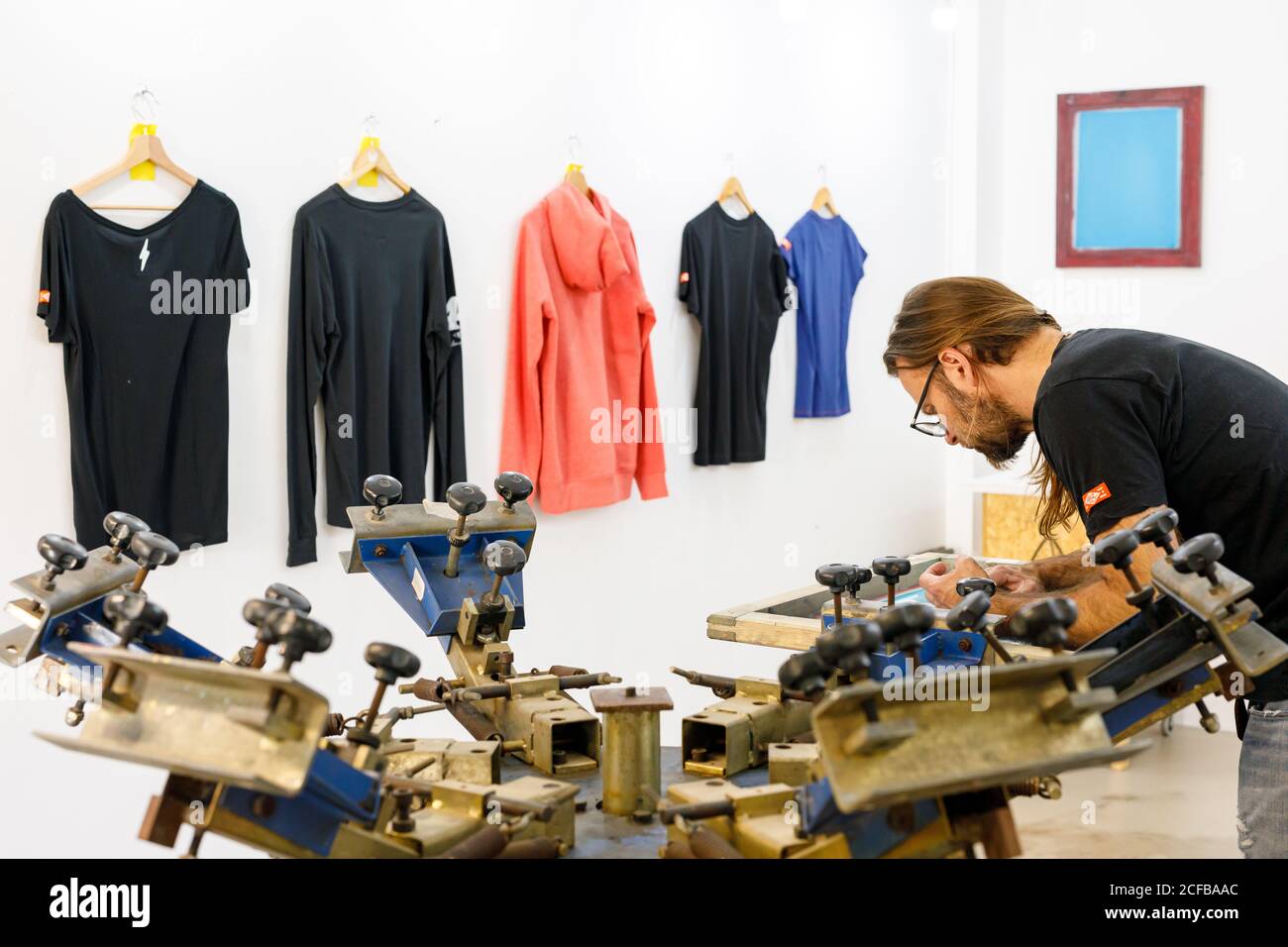 Artiste masculin concentré dans le tablier sale travaillant avec écran de soie pendant création d'imprimés sur les tee-shirts dans l'atelier Banque D'Images Artiste masculin concentré dans le tablier sale travaillant avec écran de soie pendant création d'imprimés sur les tee-shirts dans l'atelier Banque D'Images