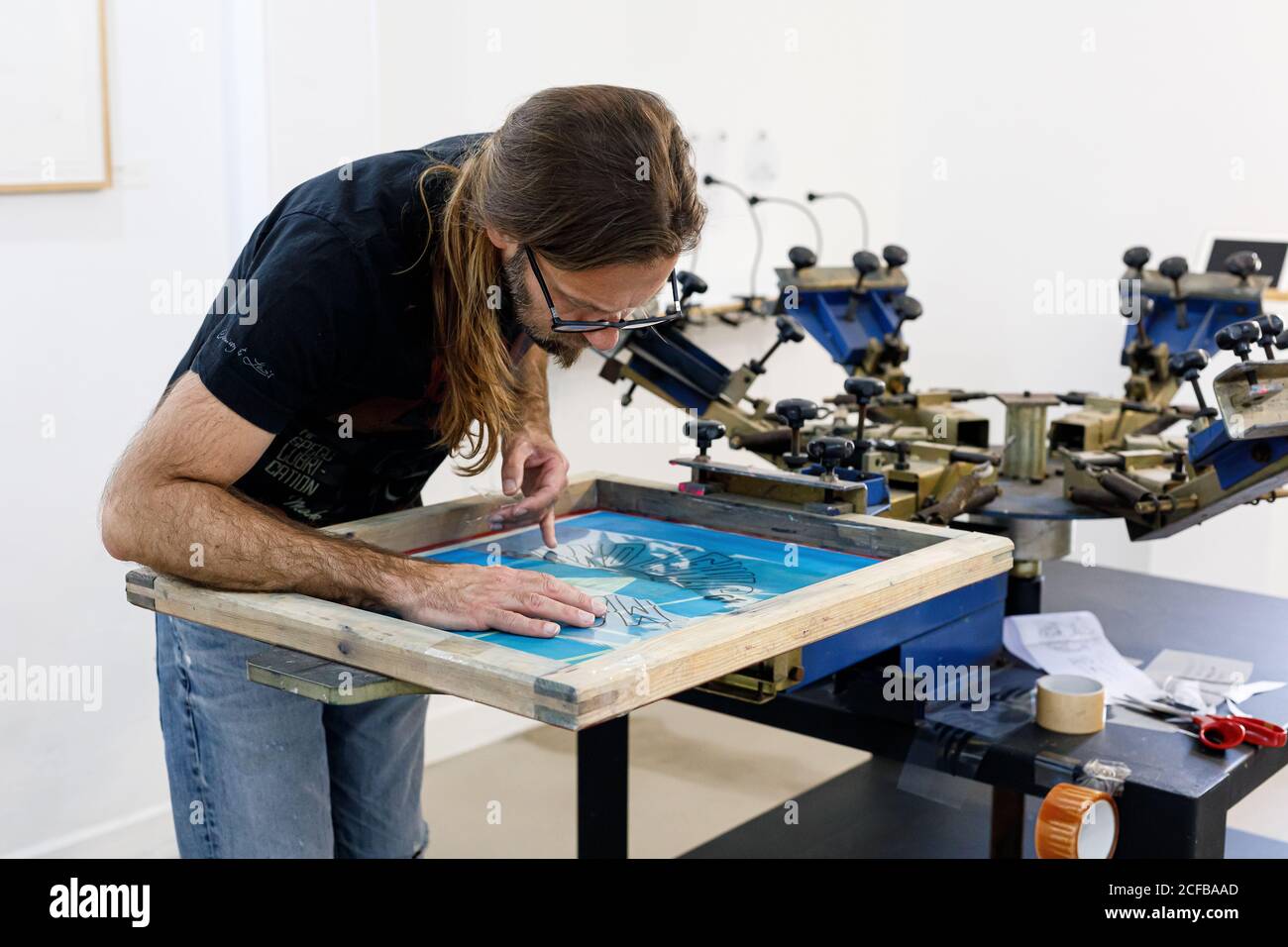 Artiste masculin concentré dans le tablier sale travaillant avec écran de soie pendant création d'imprimés sur les tee-shirts dans l'atelier Banque D'Images Artiste masculin concentré dans le tablier sale travaillant avec écran de soie pendant création d'imprimés sur les tee-shirts dans l'atelier Banque D'Images
