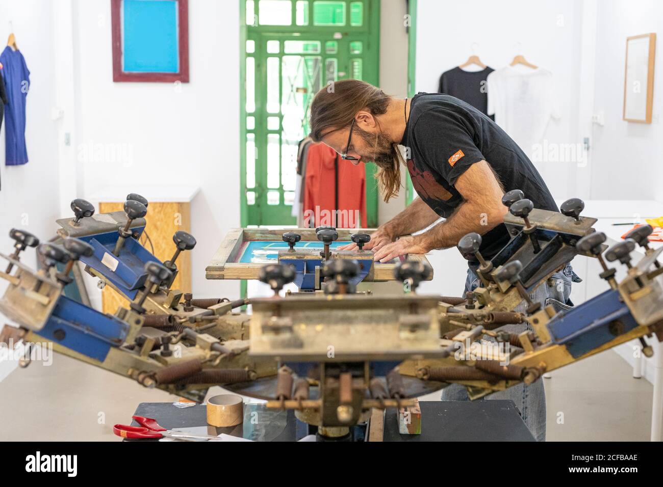 Artiste masculin concentré dans le tablier sale travaillant avec écran de soie pendant création d'imprimés sur les tee-shirts dans l'atelier Banque D'Images Artiste masculin concentré dans le tablier sale travaillant avec écran de soie pendant création d'imprimés sur les tee-shirts dans l'atelier Banque D'Images