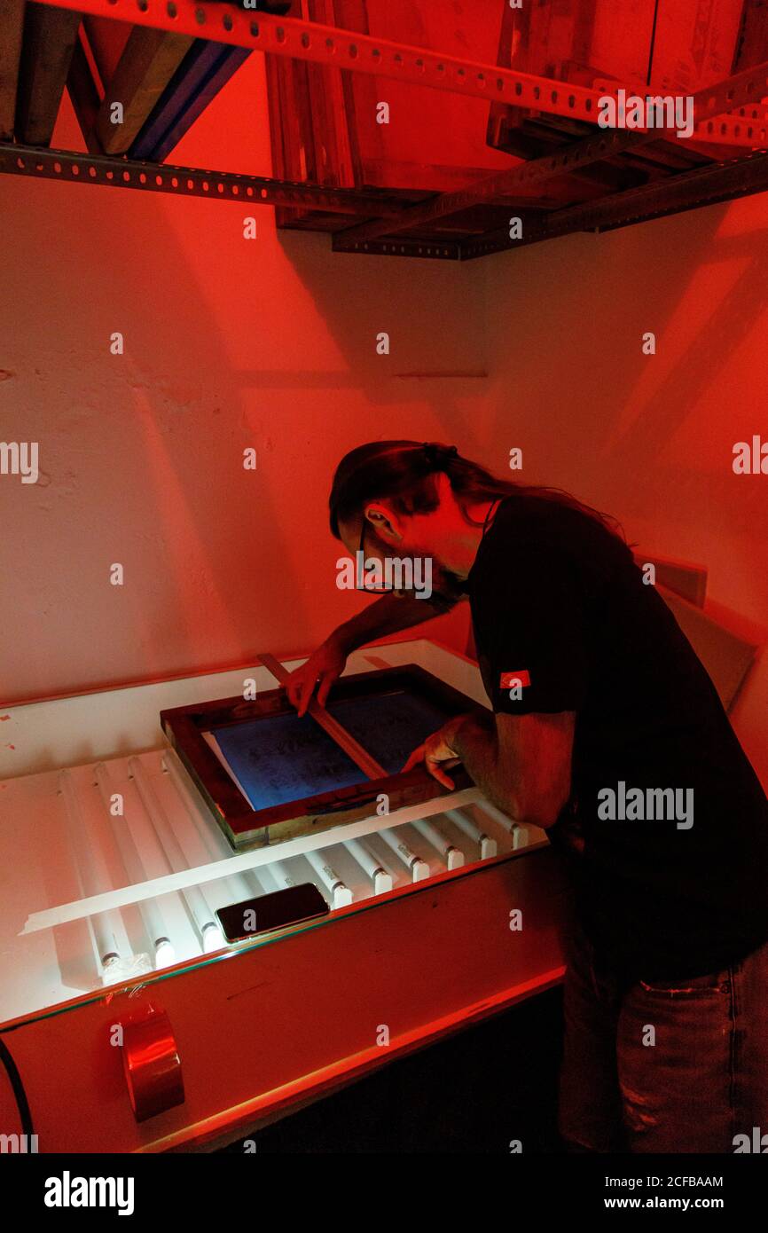 Artiste masculine concentrée avec sèche cheveux longs écran d'impression rouge salle d'atelier éclairée Banque D'Images