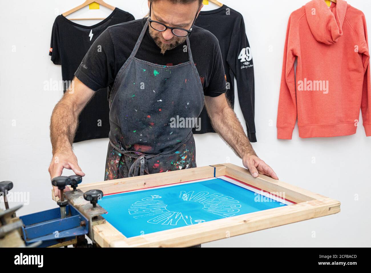 Artiste masculin concentré dans le tablier sale travaillant avec écran de soie pendant création d'imprimés sur les tee-shirts dans l'atelier Banque D'Images Artiste masculin concentré dans le tablier sale travaillant avec écran de soie pendant création d'imprimés sur les tee-shirts dans l'atelier Banque D'Images