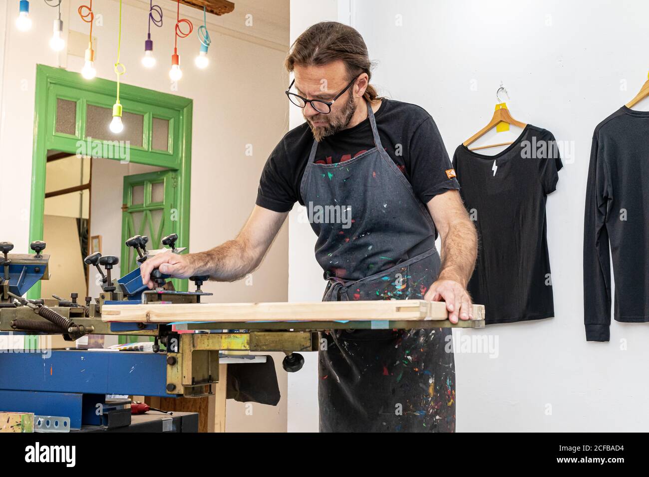 Artiste masculin concentré dans le tablier sale travaillant avec écran de soie pendant création d'imprimés sur les tee-shirts dans l'atelier Banque D'Images Artiste masculin concentré dans le tablier sale travaillant avec écran de soie pendant création d'imprimés sur les tee-shirts dans l'atelier Banque D'Images