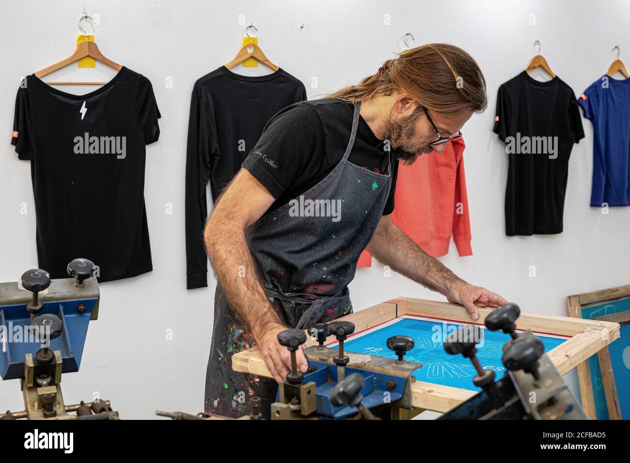 Artiste masculin concentré dans le tablier sale travaillant avec écran de soie pendant création d'imprimés sur les tee-shirts dans l'atelier Banque D'Images Artiste masculin concentré dans le tablier sale travaillant avec écran de soie pendant création d'imprimés sur les tee-shirts dans l'atelier Banque D'Images