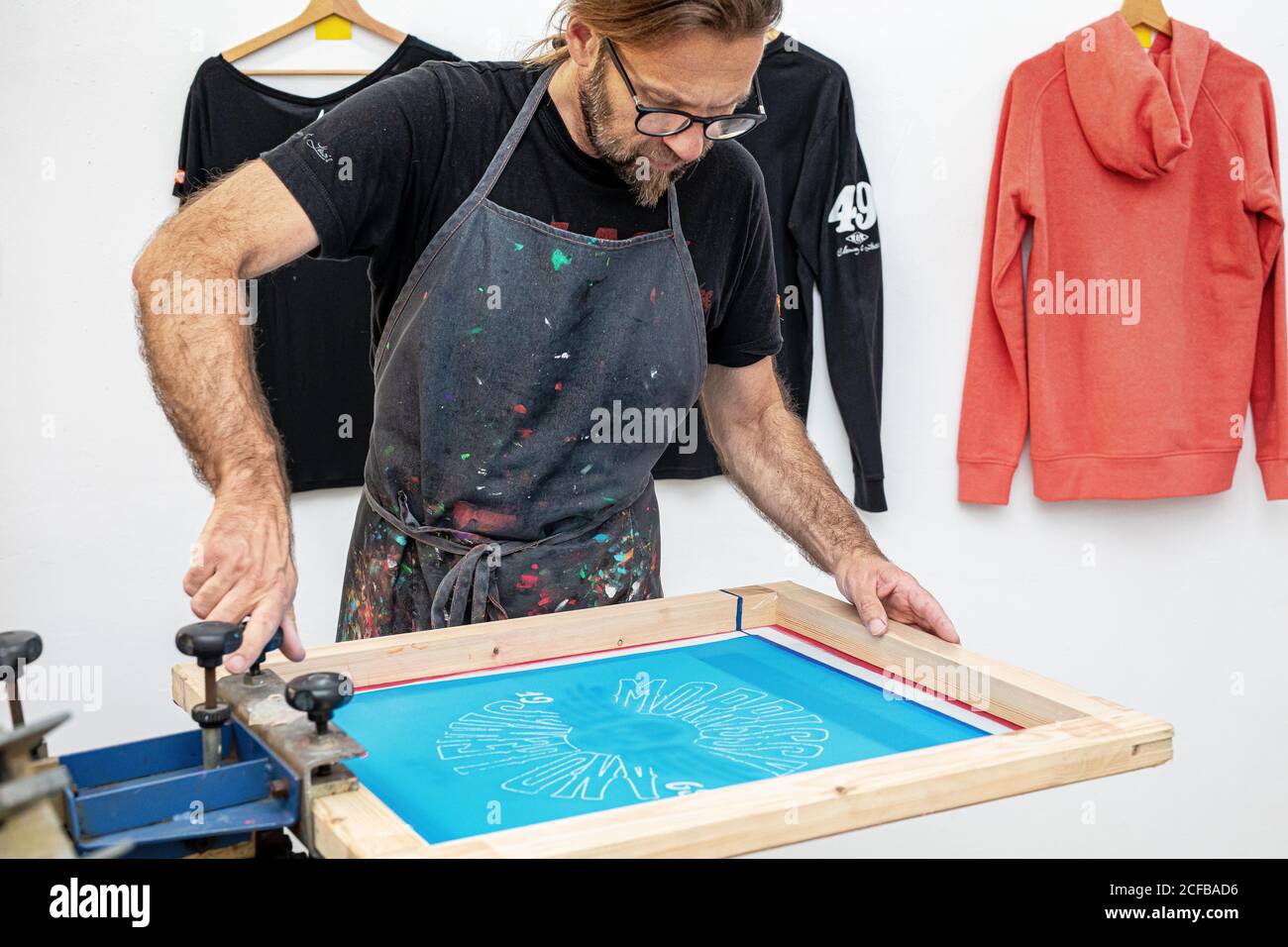 Artiste masculin concentré dans le tablier sale travaillant avec écran de soie pendant création d'imprimés sur les tee-shirts dans l'atelier Banque D'Images Artiste masculin concentré dans le tablier sale travaillant avec écran de soie pendant création d'imprimés sur les tee-shirts dans l'atelier Banque D'Images