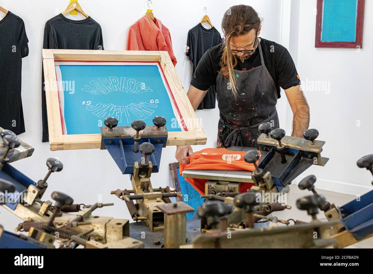 Artiste masculin concentré dans le tablier sale travaillant avec écran de soie pendant création d'imprimés sur les tee-shirts dans l'atelier Banque D'Images Artiste masculin concentré dans le tablier sale travaillant avec écran de soie pendant création d'imprimés sur les tee-shirts dans l'atelier Banque D'Images