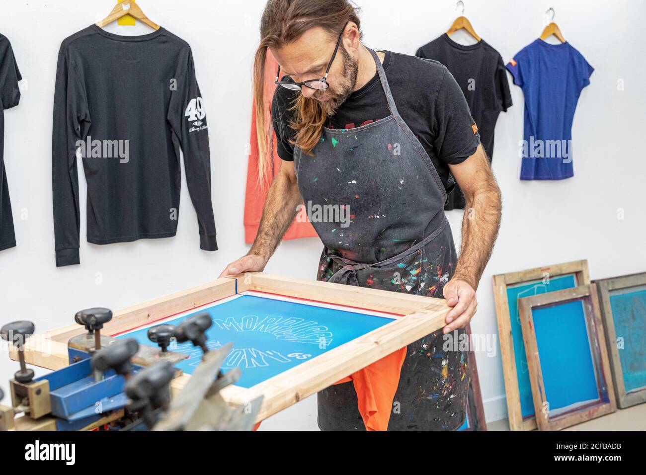 Artiste masculin concentré dans le tablier sale travaillant avec écran de soie pendant création d'imprimés sur les tee-shirts dans l'atelier Banque D'Images Artiste masculin concentré dans le tablier sale travaillant avec écran de soie pendant création d'imprimés sur les tee-shirts dans l'atelier Banque D'Images