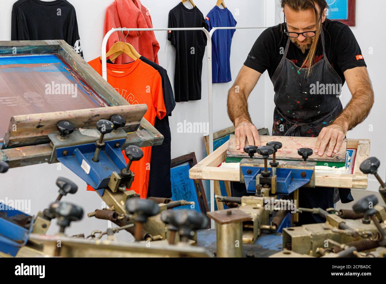 Artiste masculin concentré dans le tablier sale travaillant avec écran de soie pendant création d'imprimés sur les tee-shirts dans l'atelier Banque D'Images Artiste masculin concentré dans le tablier sale travaillant avec écran de soie pendant création d'imprimés sur les tee-shirts dans l'atelier Banque D'Images