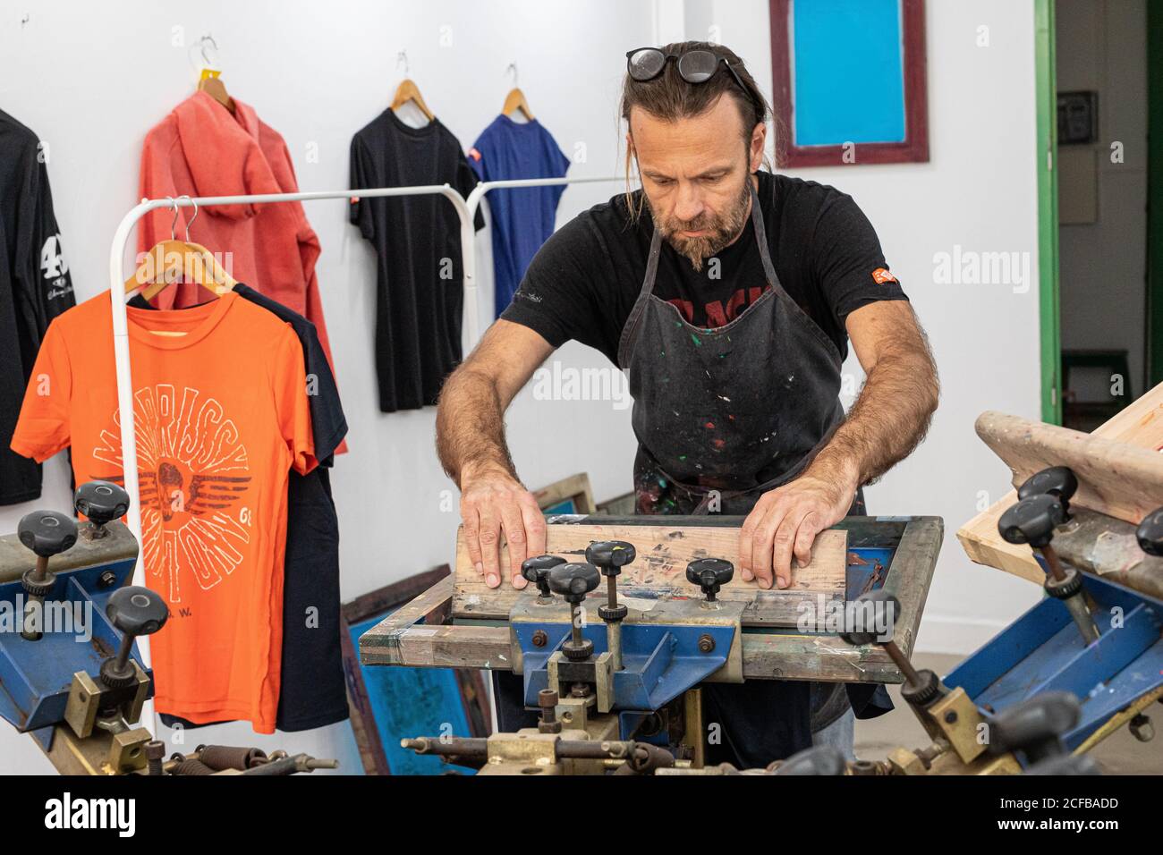 Artiste masculin concentré dans le tablier sale travaillant avec écran de soie pendant création d'imprimés sur les tee-shirts dans l'atelier Banque D'Images