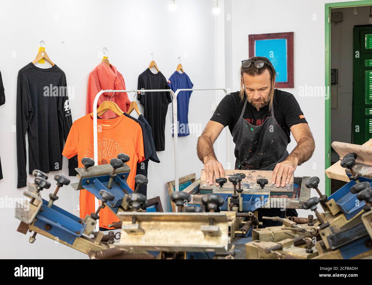 Artiste masculin concentré dans le tablier sale travaillant avec écran de soie pendant création d'imprimés sur les tee-shirts dans l'atelier Banque D'Images Artiste masculin concentré dans le tablier sale travaillant avec écran de soie pendant création d'imprimés sur les tee-shirts dans l'atelier Banque D'Images