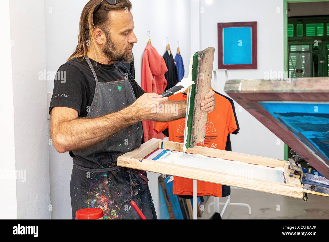 Artiste masculin concentré dans le tablier sale travaillant avec écran de soie pendant création d'imprimés sur les tee-shirts dans l'atelier Banque D'Images Artiste masculin concentré dans le tablier sale travaillant avec écran de soie pendant création d'imprimés sur les tee-shirts dans l'atelier Banque D'Images