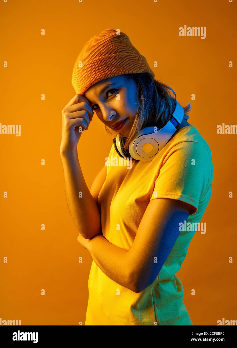 Jeune femme élégante et attentionnés avec un bonnet orange et un casque autour du cou, tête au-dessus des bras et regardant l'appareil photo sur fond orange Banque D'Images