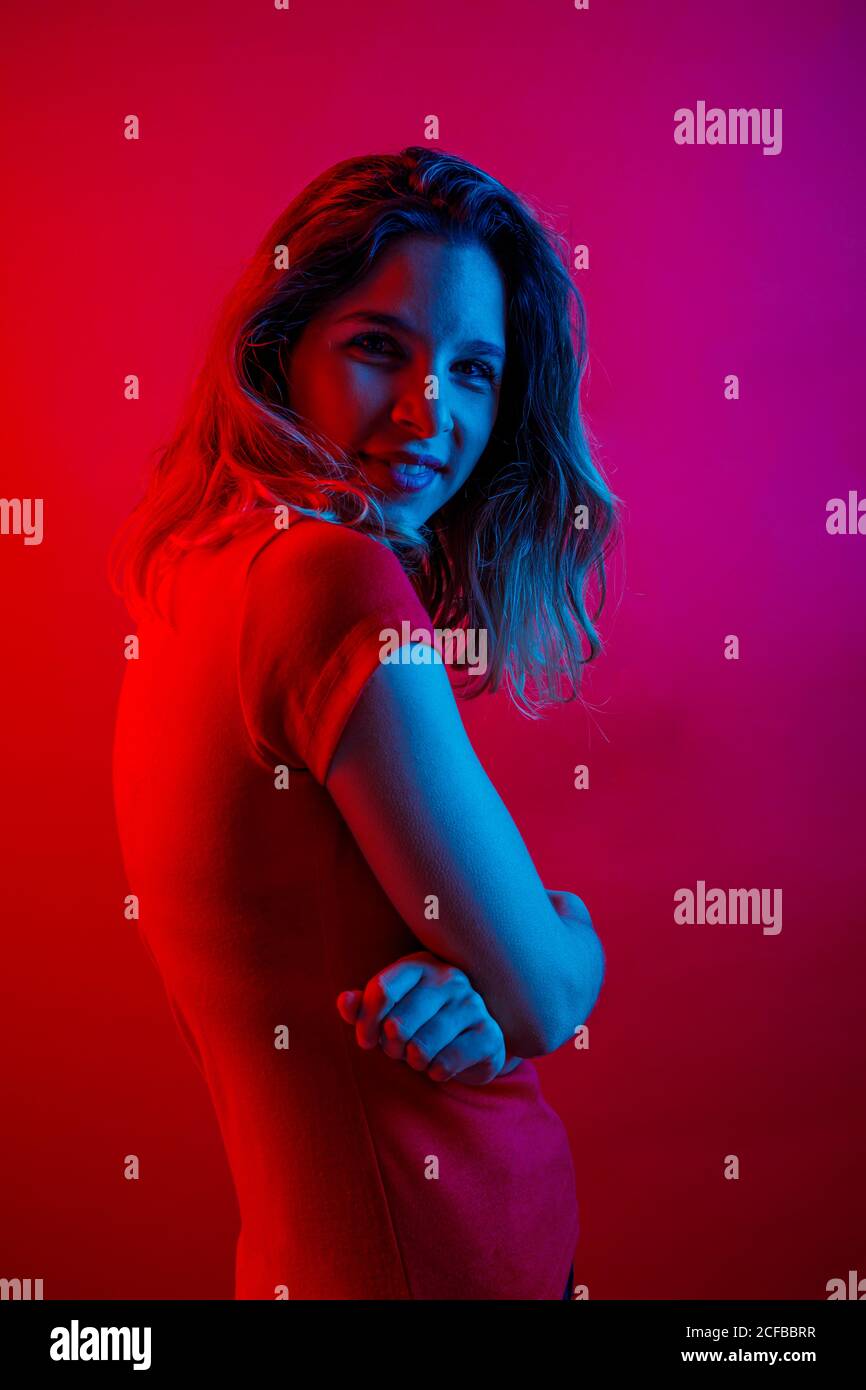 Vue latérale d'une jeune femme heureuse aux bras croisés souriant et regardant la caméra tout en se tenant sous un éclairage au néon bleu sur fond rouge Banque D'Images