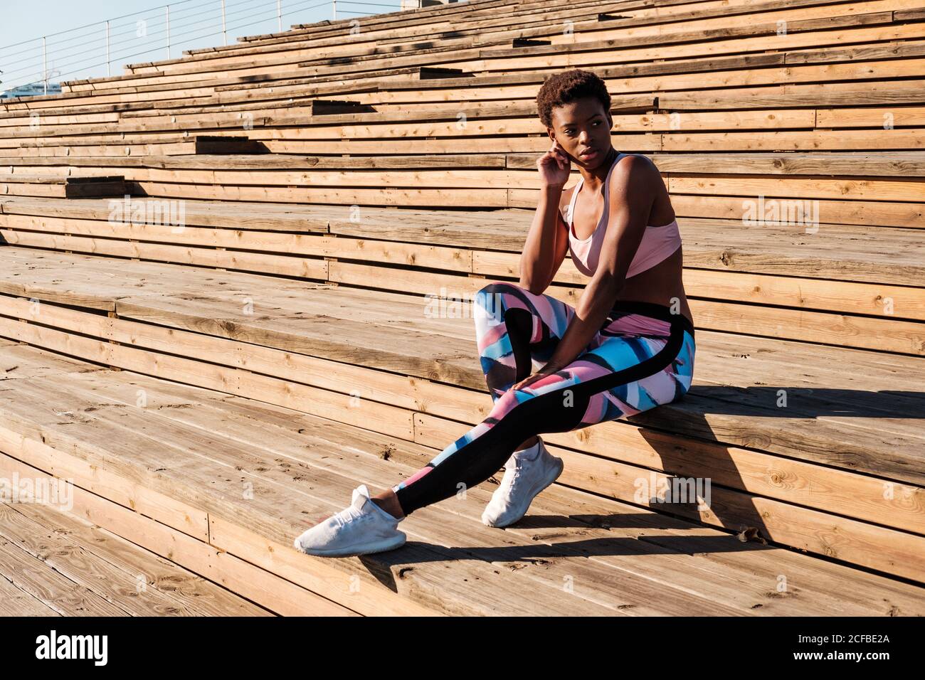 Jeune femme afro-américaine mince en vêtements de sport colorés et en  baskets blanches, regardant loin avec curiosité et contemplant tout en  étant assise seule sur des escaliers en bois marron sur le