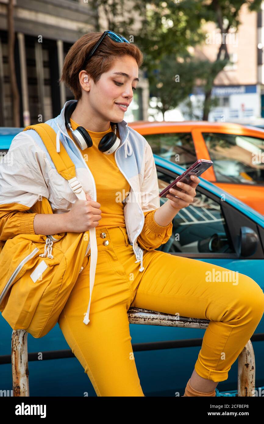 Jeune femme moderne dans des vêtements jaune tendance avec casque sans fil  et un sac à main jaune pour surfer sur votre téléphone mobile tout en étant  assis sur une clôture contre